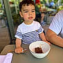 child, boy, dessert, bowl, spoon, table, striped_shirt, indoor, blurred_background, food, casual, person, sitting, young, cute, meal, cafe, daylight, happy, portrait