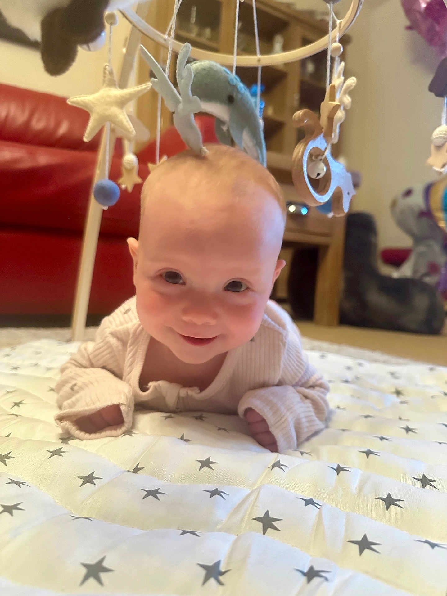 Isabel is registered to the contest to win money with this photo: baby, carpet, child, cozy, cute, face, floor, happy, home, indoors, infant, mobile, person, playful, quilt, red_couch, smiling, soft_toys, star_pattern, wooden_furniture