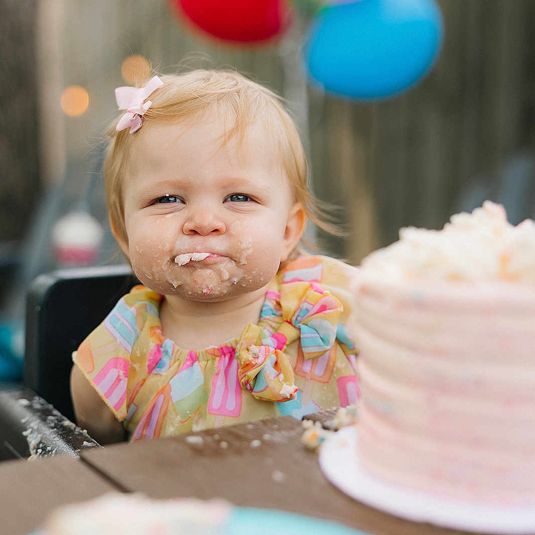 Ellyette is registered to the contest to win money with this photo: balloons, birthday, cake, celebration, child, colorful, food, frosting, fun, happy, high_chair, messy_face, outdoor, party, pink_bow, plate, portrait, smile, table, toddler