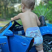 Tobias is registered to the contest to win money with this photo: toddler, diaper, toy_car, blue, outdoor, child, back_view, grass, trees, nature, footwear, side_mirror, door, wheel, curious, baby, playing, summer, sunlight, barefoot