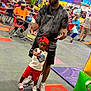 child, man, indoor, play_area, cap, red_shorts, black_shirt, sneakers, people, colorful, floor, tables, chairs, background, gesture, posing, casual_clothing, adult, kid, fun