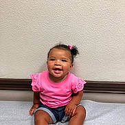 Jersey is registered to the contest to win money with this photo: baby, bright, casual_clothing, child, cute, daylight, denim_shorts, hair_bow, happy, indoor, person, pink_shirt, portrait, sitting, skin, smile, toddler, wall, white_shoes, young_child