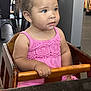 toddler, child, high_chair, pink_dress, curly_hair, blue_eyes, wooden_furniture, restaurant, indoor, casual, person, seated, hand, face, clothing, young_child, cute, portrait, table, looking_away