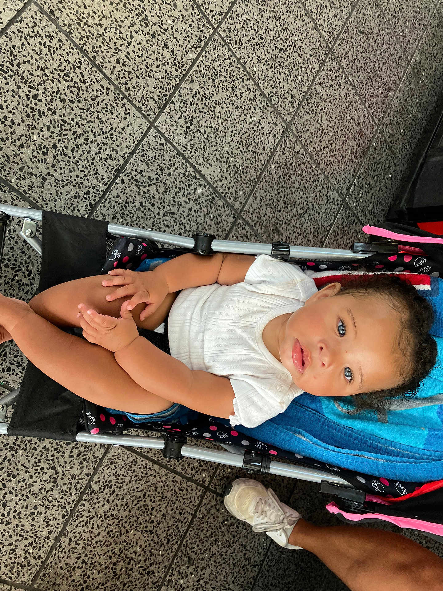 Jersey is registered to the contest to win money with this photo: toddler, child, stroller, blue_eyes, white_shirt, denim_shorts, towel, curly_hair, footwear, sneaker, floor_tiles, person, legs, hand, seated, indoor, casual_clothing, baby, infant, cute