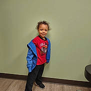 Koda is registered to the contest to win money with this photo: child, smiling, curly_hair, red_shirt, blue_jacket, black_pants, sneakers, standing, indoor, wooden_floor, green_wall, office_chair, casual_clothing, person, portrait, happy, young, full_body, confident, wall