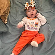 Lucia is registered to the contest to win money with this photo: baby, child, infant, halloween, costume, orange_pants, headband, bow, bed, blanket, white_shirt, text_on_clothing, wide_eyes, cute, lying_down, sock, indoors, person, celebration, holiday