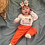 baby, child, infant, halloween, costume, orange_pants, headband, bow, bed, blanket, white_shirt, text_on_clothing, wide_eyes, cute, lying_down, sock, indoors, person, celebration, holiday