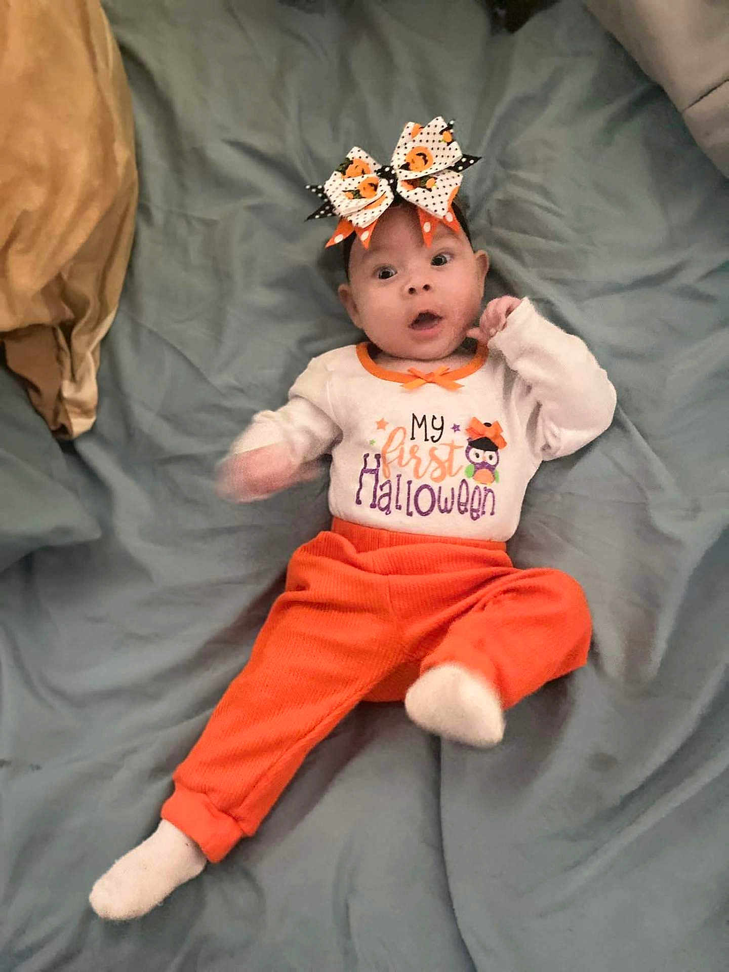 Lucia is registered to the contest to win money with this photo: baby, child, infant, halloween, costume, orange_pants, headband, bow, bed, blanket, white_shirt, text_on_clothing, wide_eyes, cute, lying_down, sock, indoors, person, celebration, holiday