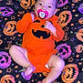 baby, black_background, blanket, child, cute, face, feet, halloween_theme, hands, indoor, infant, legs, lying_down, orange_clothing, pacifier, pattern, portrait, pumpkin_outfit, purple, stars