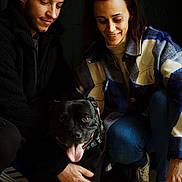 Reïko participe au concours pour gagner de l'argent avec cette photo : dog, black_dog, pet, smiling, person, man, woman, crouching, indoor, casual_clothing, jeans, sneakers, jacket, collar, tongue_out, happy, portrait, friendship, companionship, warmth