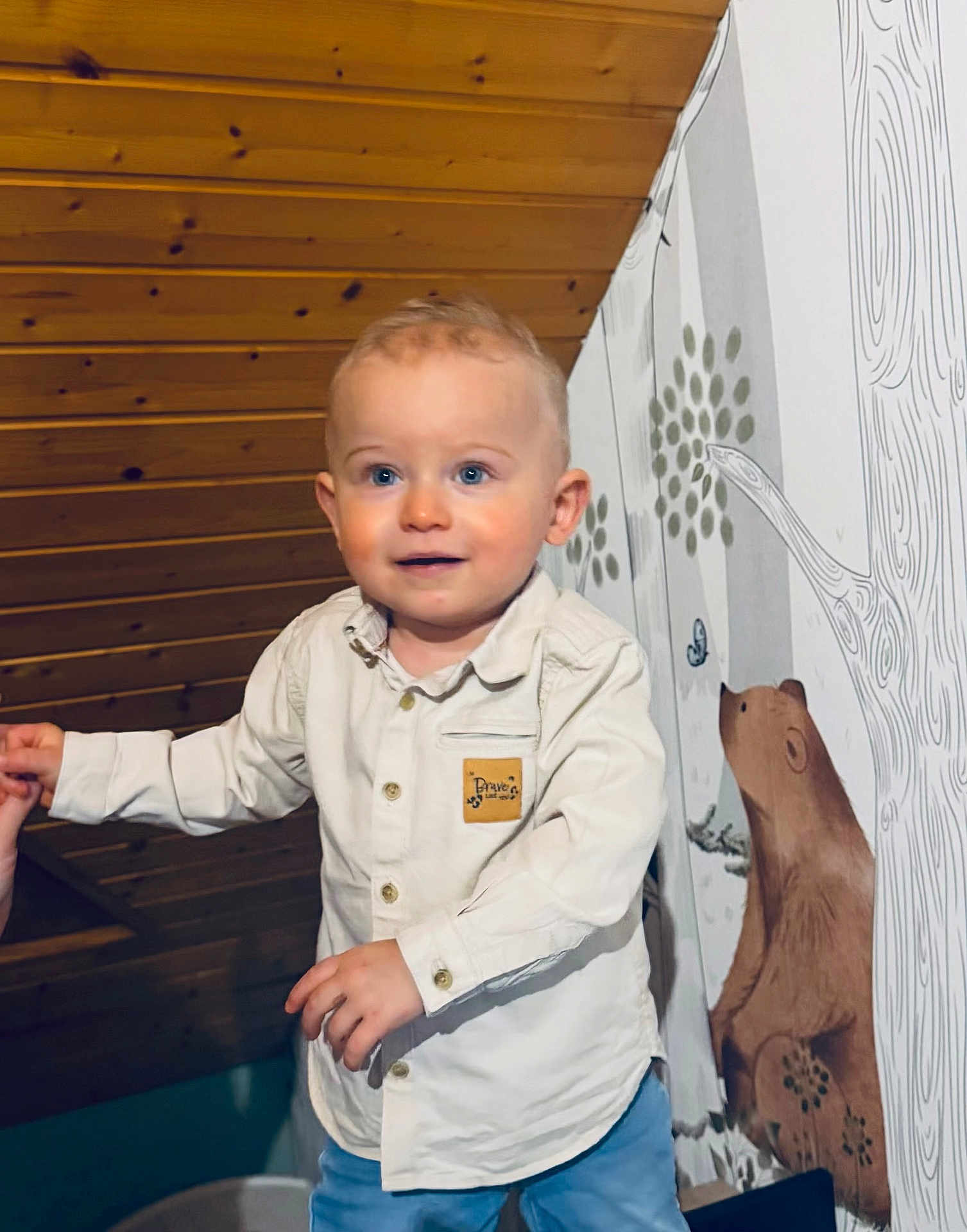 Pierre participe au concours pour gagner de l'argent avec cette photo : toddler, child, indoor, portrait, smile, blue_eyes, white_shirt, blue_pants, wooden_ceiling, wall_mural, bear, hand_holding, curly_hair, cute, happy, person, baby, clothing, standing, room