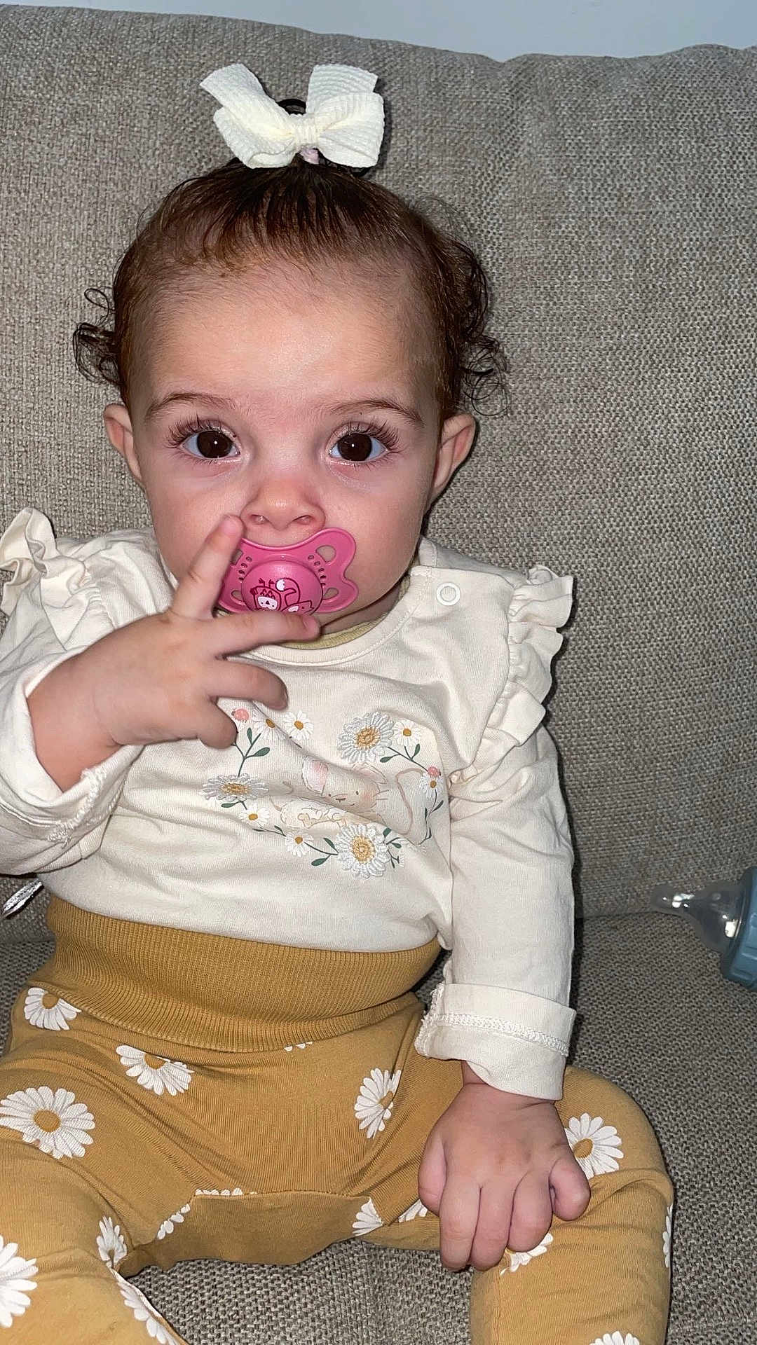 Mahira a rejoint le concours — aidez-le/la à gagner de superbes lots ! baby, child, pacifier, bow, couch, clothing, person, sitting, hand, face, eyes, cute, infant, patterned_pants, floral, yellow, white, pink, portrait, indoor