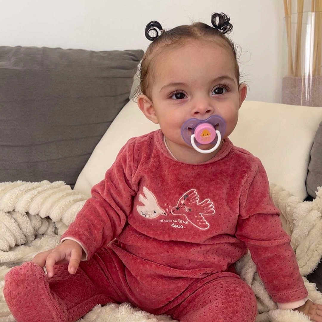 Mahira participe au concours pour gagner de l'argent avec cette photo : baby_clothes, blanket, child, couch, cozy, cute, face, hair_buns, happy, home, indoors, pacifier, person, portrait, red_outfit, sitting, smiling, soft_texture, toddler, young_child