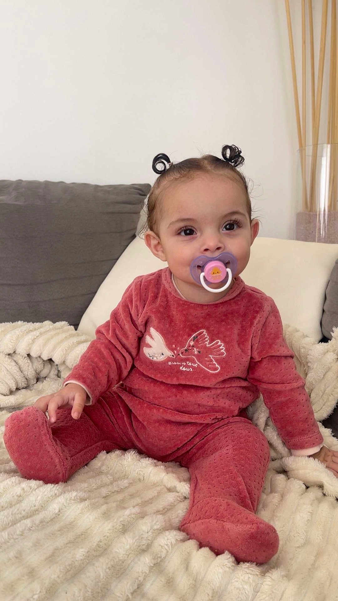 Mahira participe au concours pour gagner de l'argent avec cette photo : baby_clothes, blanket, child, couch, cozy, cute, face, hair_buns, happy, home, indoors, pacifier, person, portrait, red_outfit, sitting, smiling, soft_texture, toddler, young_child