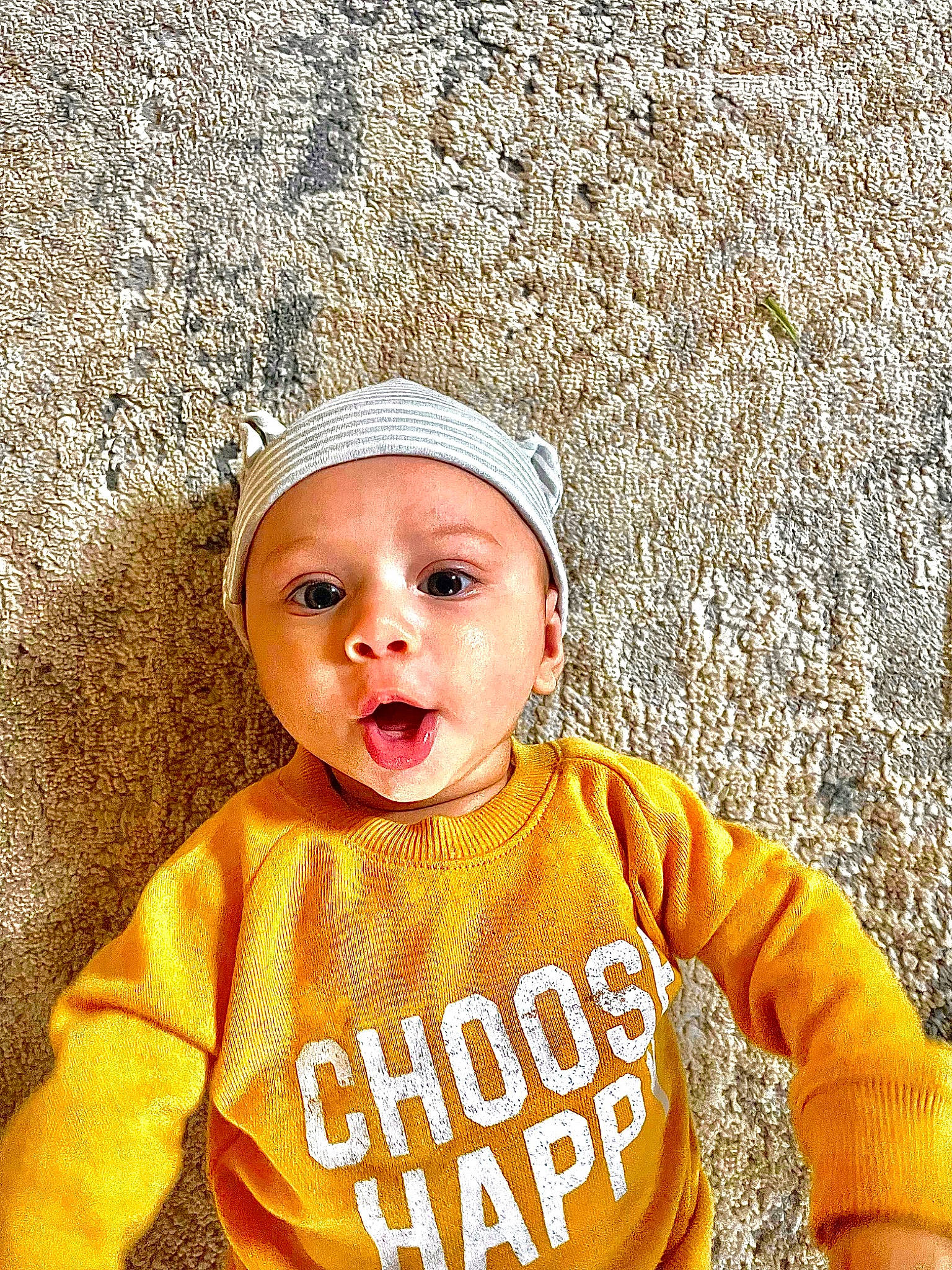 Callum is registered to the contest to win money with this photo: baby, baby_toddler_clothing, beanie, cap, child, cool, eye, face, fashion_accessory, fun, happy, hat, headwear, hood, outerwear, person, plant, sitting, sleeve, street_fashion