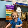 Matthew is registered to the contest to win money with this photo: blue_clothing, chair, child, classroom, drawer, educational_setting, floor, furniture, happy, indoor, person, plastic_toy, playful, smiling, stacked_chairs, stacked_drawers, toddler, toy, wooden_floor, young_child