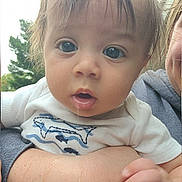 Wyatt is registered to the contest to win money with this photo: baby, child, infant, portrait, close_up, face, wide_eyes, looking_at_camera, drooling, open_mouth, blonde_hair, white_shirt, clothing, adult, parent, being_held, hand, arm, outdoor, trees