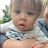 baby, child, infant, portrait, close_up, face, wide_eyes, looking_at_camera, drooling, open_mouth, blonde_hair, white_shirt, clothing, adult, parent, being_held, hand, arm, outdoor, trees
