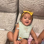 Meilani joined the competition — help win amazing prizes! baby, child, infant, couch, sofa, blanket, bib, headband, yellow_bow, holding_hands, smiling, happy, indoor, person, seated, furniture, textile, skin, foot, hand