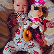 Kiana is registered to the contest to win money with this photo: baby, carpet, child, comfort, cushion, cute, face, footwear, hand, indoors, infant, lying_down, minnie_mouse, pajamas, person, plush_toy, smile, snowman_pattern, soft_toy, toy