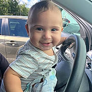 Lukai is registered to the contest to win money with this photo: baby, child, smiling, car_interior, steering_wheel, driver_seat, clothing, short_sleeves, blanket, hand, face, eyes, happy, cute, daylight, window, outdoor, vehicle, seat, portrait