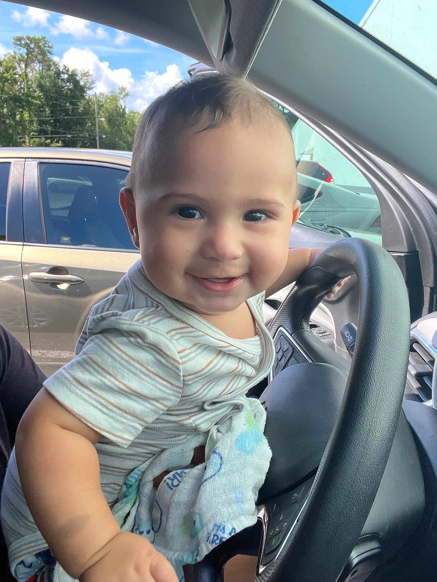 Lukai is registered to the contest to win money with this photo: baby, child, smiling, car_interior, steering_wheel, driver_seat, clothing, short_sleeves, blanket, hand, face, eyes, happy, cute, daylight, window, outdoor, vehicle, seat, portrait