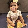 baby, bracelet, child, closeup, curly_hair, cute, face, happy, headband, indoors, infant, onesie, person, playful, portrait, sitting, smile, yellow_bow, yellow_socks, young_child