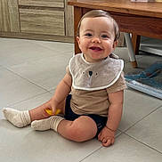 Milo a rejoint le concours — aidez-le/la à gagner de superbes lots ! baby, child, smiling, indoor, floor, toy, bib, socks, tiled_floor, table, wooden_furniture, shorts, happy, cute, person, small_child, cabinets, home, seated, playful