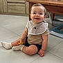 baby, child, smiling, indoor, floor, toy, bib, socks, tiled_floor, table, wooden_furniture, shorts, happy, cute, person, small_child, cabinets, home, seated, playful