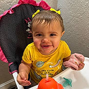 Kylie is registered to the contest to win money with this photo: toddler, child, smiling, high_chair, yellow_shirt, butterfly_print, pink_mitten, sippy_cup, pacifier, minnie_mouse, indoor, happy, cute, person, face, baby, hair_bows, eyes, mouth, hand
