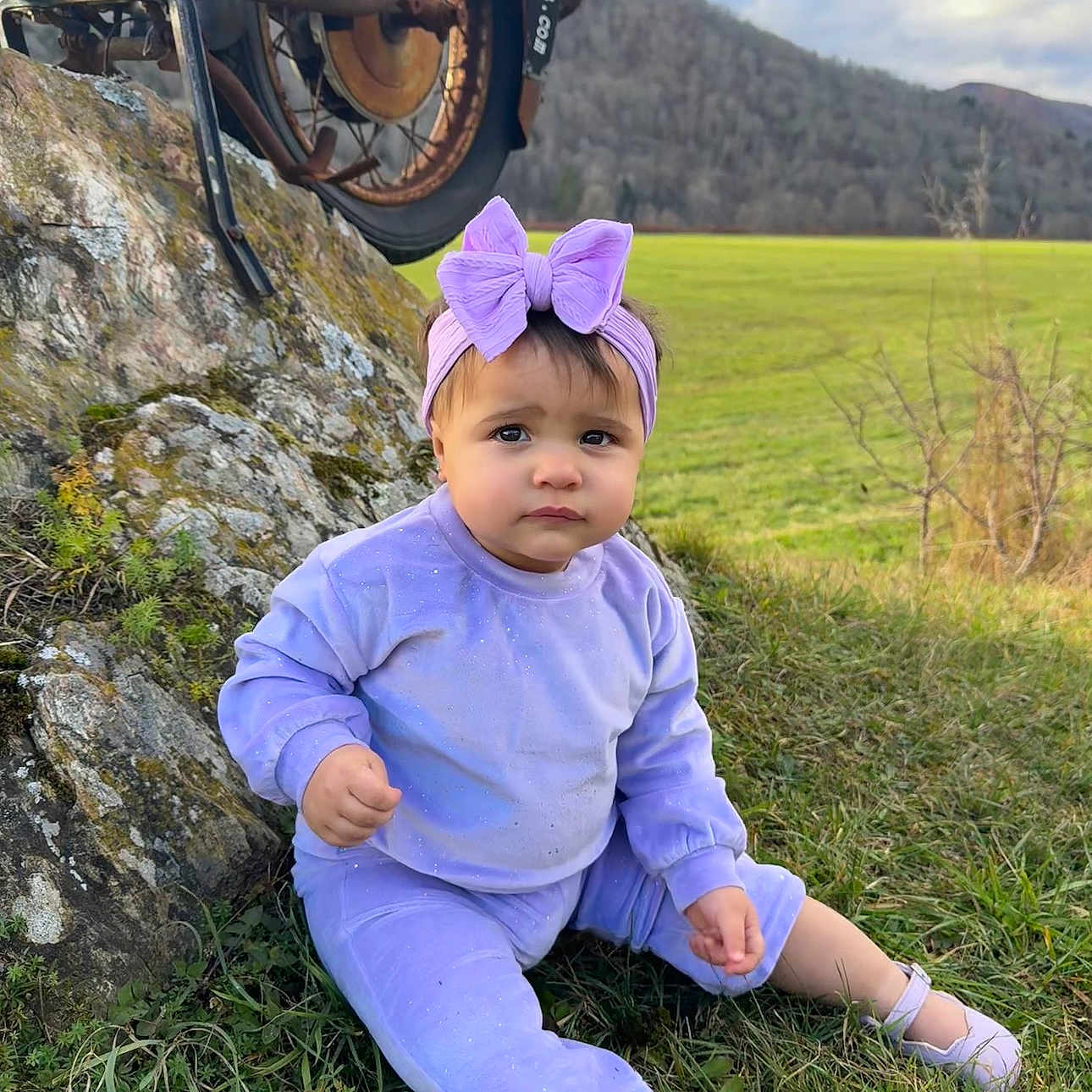 Kylie joined the competition — help win amazing prizes! bow, casual, child, cloudy_sky, cute, field, grass, headband, motorcycle_wheel, mountain, nature, outdoor, portrait, purple_clothing, rock, scenic, shoes, sitting, sparkle, toddler
