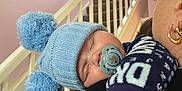 Fin is registered to the contest to win money with this photo: baby, sleeping, blue_hat, pom_poms, pacifier, adult, shoulder, staircase, hand, knitwear, earrings, indoor, pastel_wall, cozy, child, person, cuddling, warm_clothing, resting, comfort