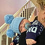 baby, sleeping, blue_hat, pom_poms, pacifier, adult, shoulder, staircase, hand, knitwear, earrings, indoor, pastel_wall, cozy, child, person, cuddling, warm_clothing, resting, comfort