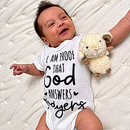 David is registered to the contest to win money with this photo: baby, infant, smiling, happy, portrait, lying_down, bed, quilt, white_bedding, plush_toy, lamb, stuffed_animal, onesie, clothing, arms, legs, dark_hair, indoor, close_up, nursery