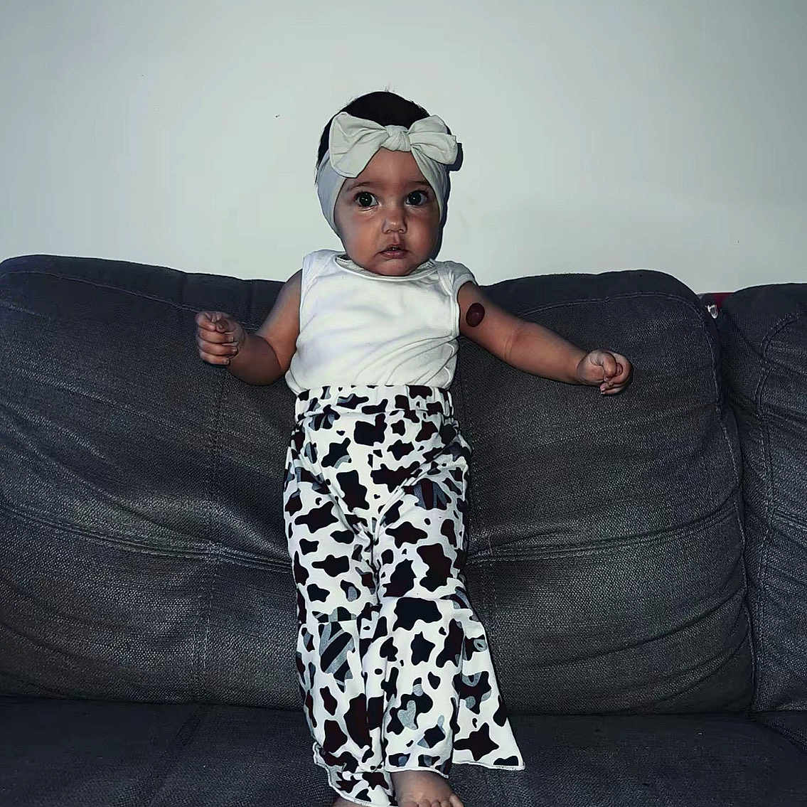 Zyaria is registered to the contest to win money with this photo: barefoot, bow, child, clothing, couch, cow_print_pants, cute, dark_background, fashion, furniture, headband, home, indoor, person, portrait, serious_expression, standing, toddler, white_top, young_child