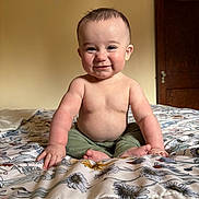 Xavier is registered to the contest to win money with this photo: baby, child, smile, sitting, bed, bedspread, floral_pattern, indoor, room, wooden_door, wall, cute, cheeks, hands, feet, bare_chest, pants, portrait, infant, happy