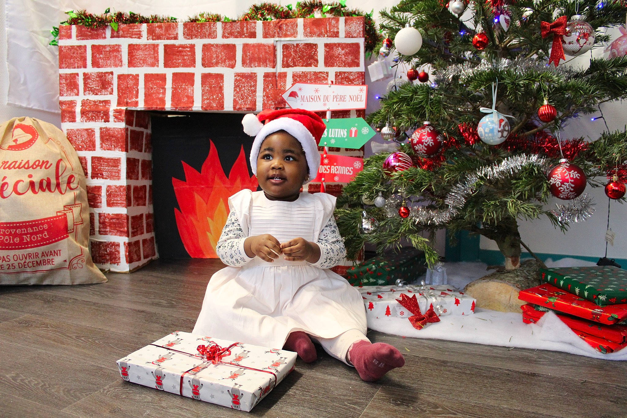 Aliyah a rejoint le concours — aidez-le/la à gagner de superbes lots ! carmine, christmas, christmas_decoration, christmas_eve, christmas_ornament, christmas_tree, event, flooring, green, happy, headwear, holiday, holiday_ornament, lap, ornament, person, red, room, smile, toddler