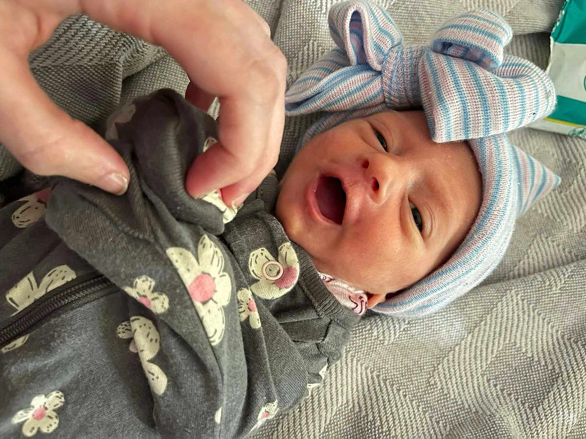 Theda is registered to the contest to win money with this photo: baby, infant, person, hand, clothing, hat, bow, floral_pattern, fabric, closeup, face, smile, newborn, cute, indoors, blanket, comfort, soft, skin, portrait