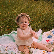 Quinn is registered to the contest to win money with this photo: baby, child, flower_crown, quilt, meadow, wildflowers, grass, outdoor, nature, smiling, clapping, summer, cute, sitting, infant, greenery, sunlight, portrait, happy, soft_light