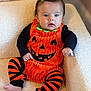 baby, infant, pumpkin_costume, halloween, orange, black, striped_leggings, green_bow, cute, child, seated, indoor, cozy, soft_texture, plush, portrait, person, furniture, armchair, expression