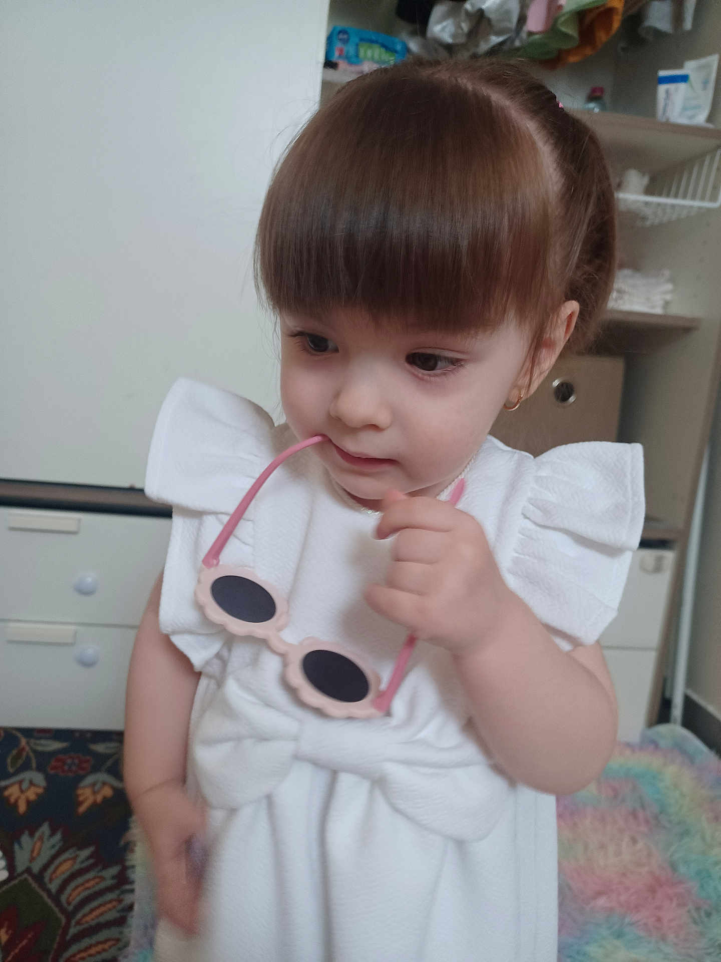 Ariana a rejoint le concours — aidez-le/la à gagner de superbes lots ! toddler, child, girl, white_dress, sunglasses, indoor, curly_hair, puffy_sleeves, cute, curious, face, hand, carpet, furniture, room, shelf, toy, earrings, portrait, soft_lighting