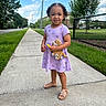 toddler, child, sidewalk, stuffed_toy, purple_dress, butterflies, sandals, smiling, curly_hair, outdoor, daytime, grass, trees, fence, suburban, person, happy, toy, summer, blue_sky