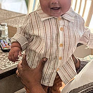 Izzak joined the competition — help win amazing prizes! baby, child, smiling, indoor, striped_shirt, brown_cap, table, chair, adult_hands, tattoo, happy, person, clothing, portrait, infant, holding, furniture, home, cute, casual
