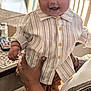 baby, child, smiling, indoor, striped_shirt, brown_cap, table, chair, adult_hands, tattoo, happy, person, clothing, portrait, infant, holding, furniture, home, cute, casual
