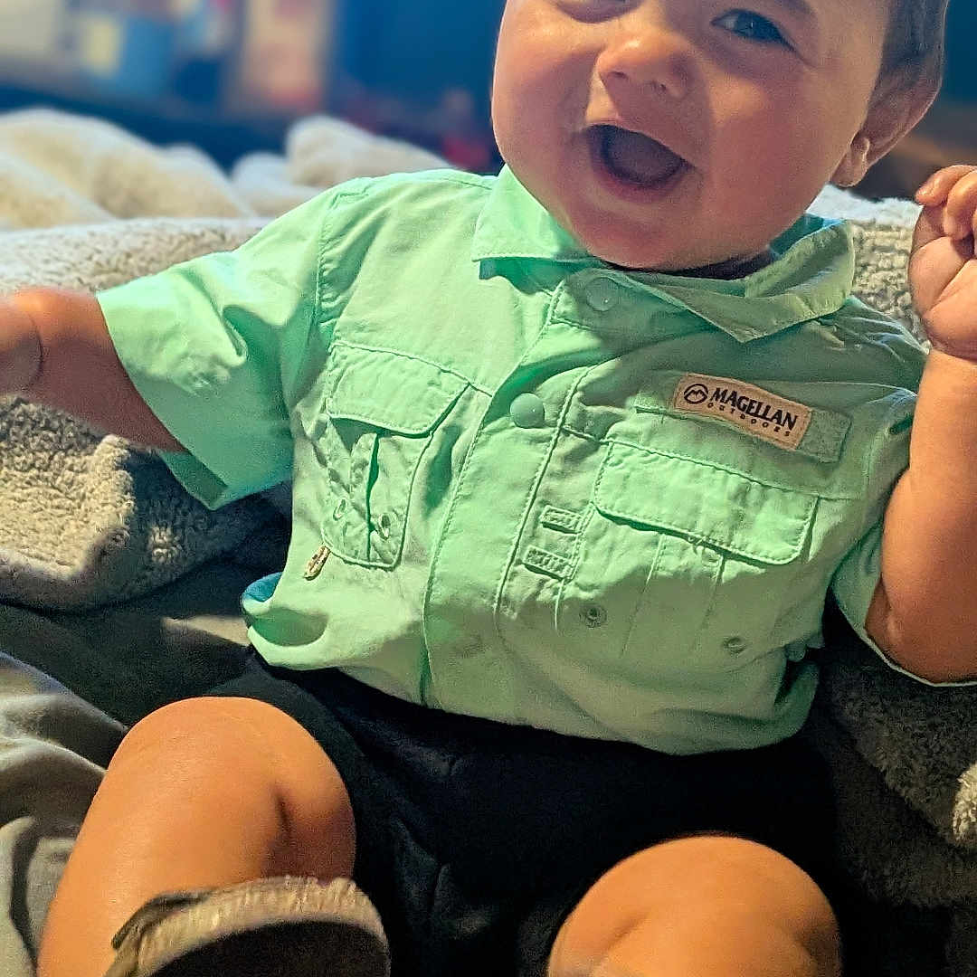 Izzak is registered to the contest to win money with this photo: baby, blanket, boots, child, clothing, cozy, cute, face, green_shirt, happy, indoor, infant, joyful, person, portrait, shirt, sitting, smiling, toddler, warm_lighting