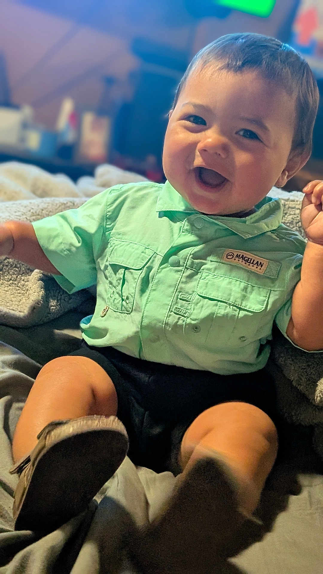 Izzak is registered to the contest to win money with this photo: baby, child, smiling, happy, shirt, green_shirt, boots, sitting, indoor, blanket, cute, portrait, infant, face, clothing, person, cozy, joyful, toddler, warm_lighting