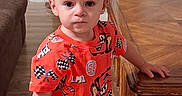 David is registered to the contest to win money with this photo: bottle, cars_theme, cartoon_print, casual, child, couch, floor, holding_object, home, indoor, living_room, person, red_clothing, serious_expression, short_hair, standing, table, toddler, wooden_furniture, young_child
