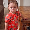 bottle, cars_theme, cartoon_print, casual, child, couch, floor, holding_object, home, indoor, living_room, person, red_clothing, serious_expression, short_hair, standing, table, toddler, wooden_furniture, young_child