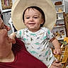 David is registered to the contest to win money with this photo: adult, basket, child, cowboy_hat, dinosaur_print, family, furniture, happy, hat, holding, home, indoor, onesie, painting, person, portrait, rocking_chair, shelf, smile, toddler