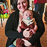 woman, baby, indoor, carpet, smile, pacifier, bib, clothing, holding, person, family, toy, furniture, jeans, black_sweater, red_top, background, home, happy, portrait